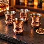 AI-generated lifestyle illustration of four Silver One handleless copper-finish shot cups with small amber tasting pours on a slate bar surface.