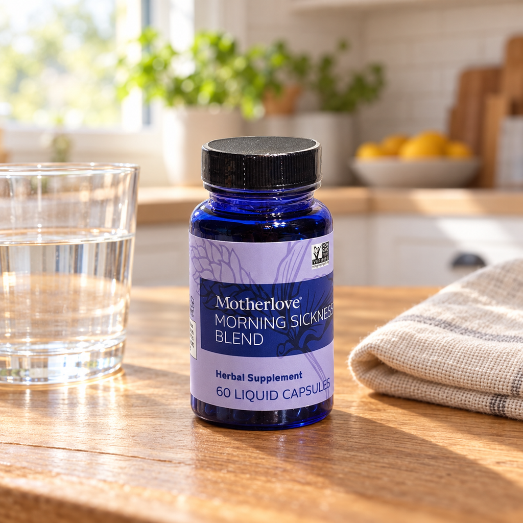 Motherlove Morning Sickness Blend 60-capsule bottle on an oak table beside water and neutral linen.