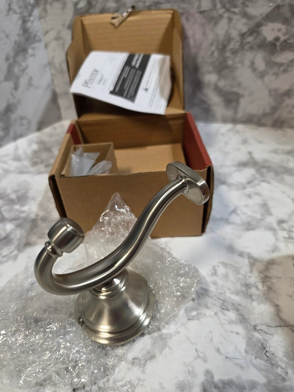 Metallic towel hook on a marble wall with a cardboard box and bubble wrap in the background.