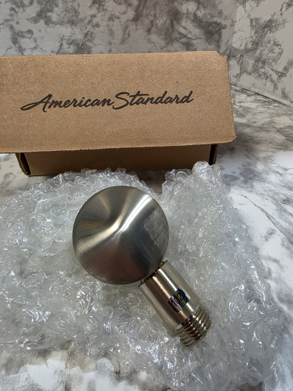 Metallic shower handle with packaging labeled 'American Standard' on a marble surface.
