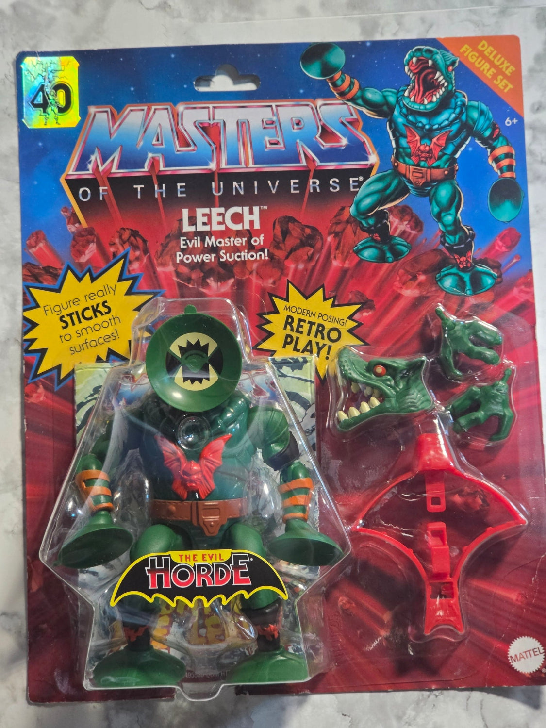 Toy figure in packaging with 'Masters of the Universe' branding on a marbled surface.