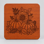 A square wooden coaster with rounded corners, held in a hand. The coaster features a laser-engraved autumn design of pumpkins, sunflowers, and leaves.