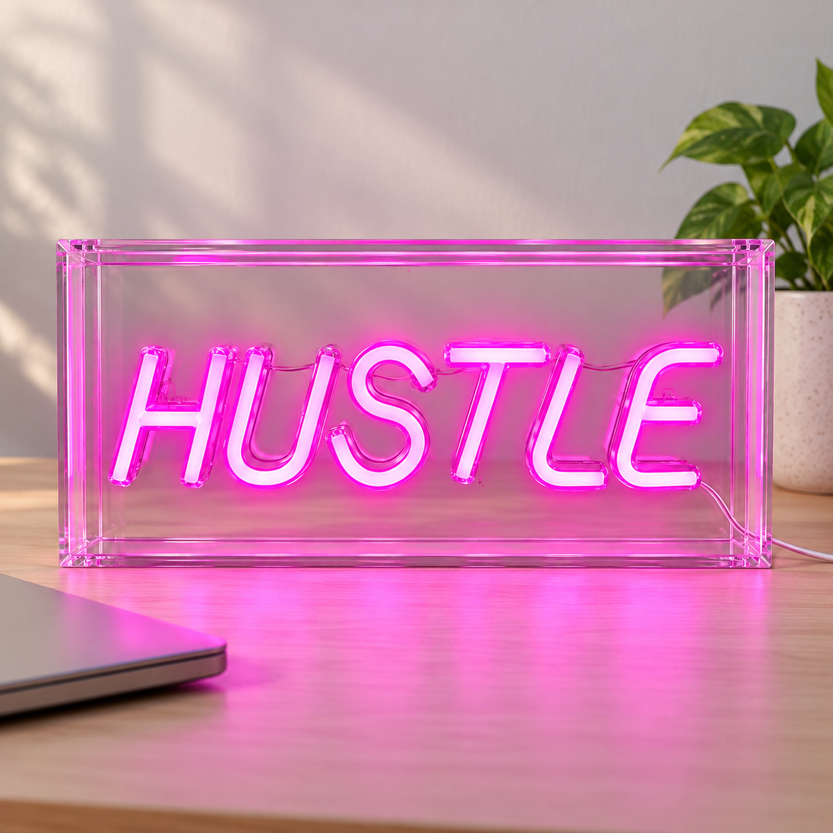 Elements HUSTLE pink LED sign on a wood desk beside a laptop and plant