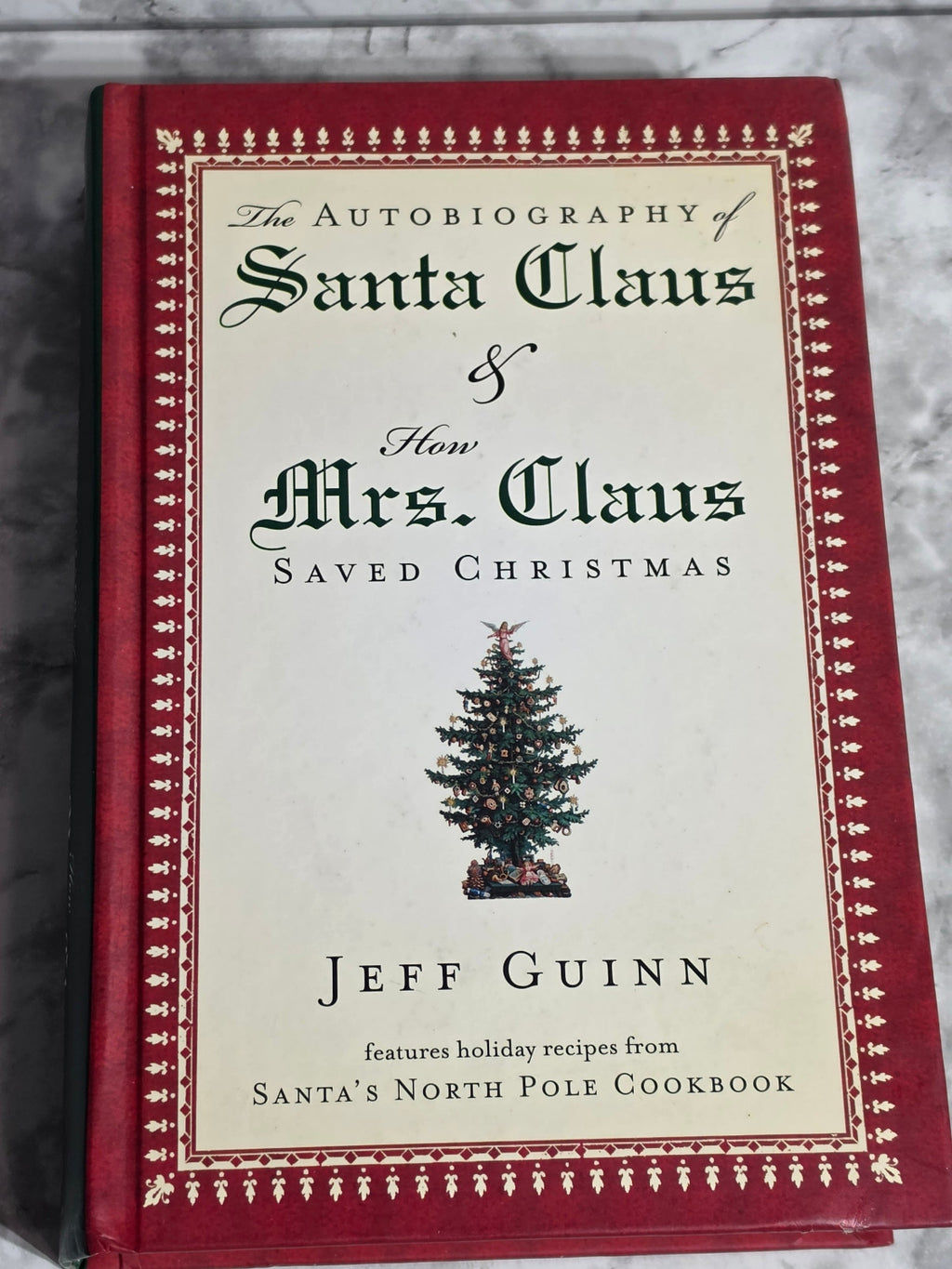 Book titled 'The Autobiography of Santa Claus & Mrs. Claus How Christmas Saved Christmas' by Jeff Guinn on a marble surface.