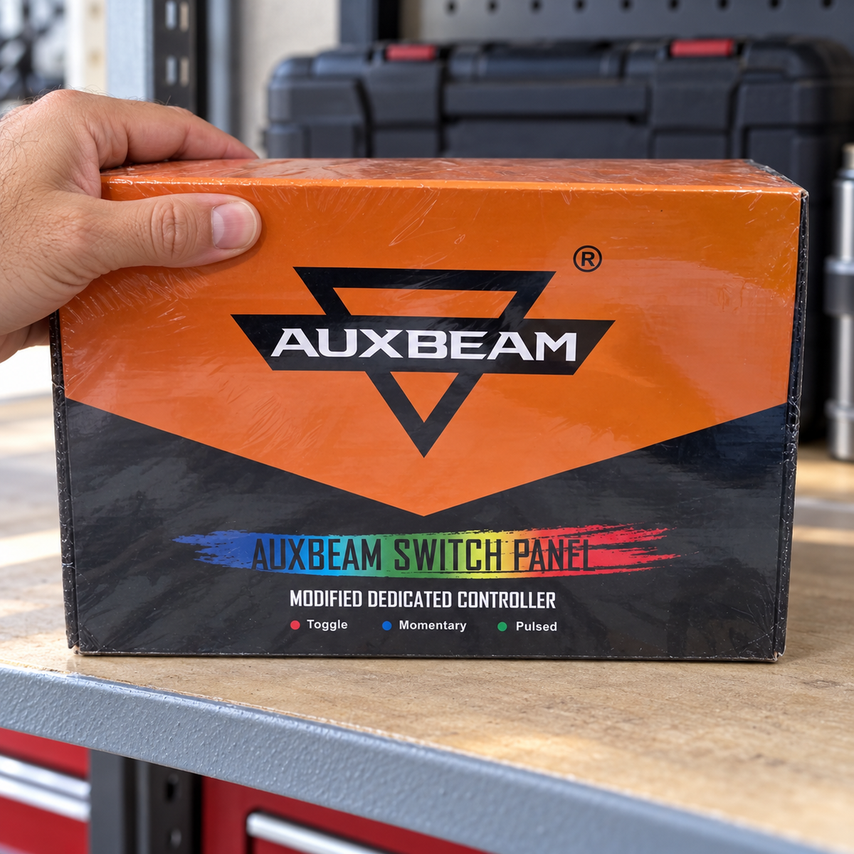 AI-generated lifestyle illustration: adult hand steadying the closed Auxbeam box in a garage work area; small label covered, contents concealed, props not included.