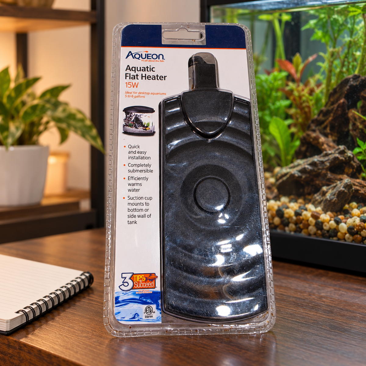 AI-generated lifestyle scene: packaged Aqueon 15W flat heater beside a notebook and planted aquarium; props not included.