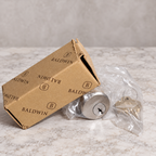 AI illustration: Baldwin 8321.150 satin nickel cylinder in clear packaging beside a worn Baldwin carton on pale stone