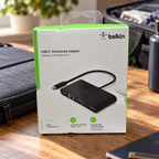 Belkin AVC005 USB-C multimedia adapter retail box in a travel packing setting, AI-assisted lifestyle illustration.