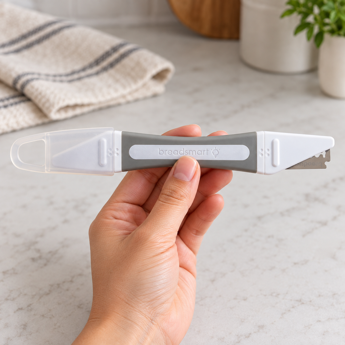 An adult hand holds a gray Breadsmart dual-ended bread lame by its center handle, with a translucent cap at one end and an exposed scoring blade at the other.