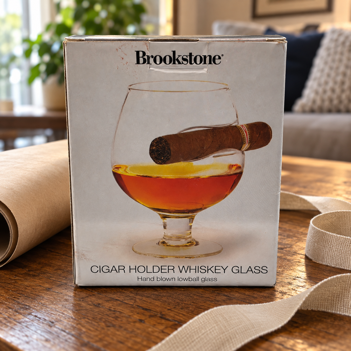 AI-created lifestyle illustration of the worn Brookstone whiskey-glass retail package beside gift paper and ribbon; wrapping props not included