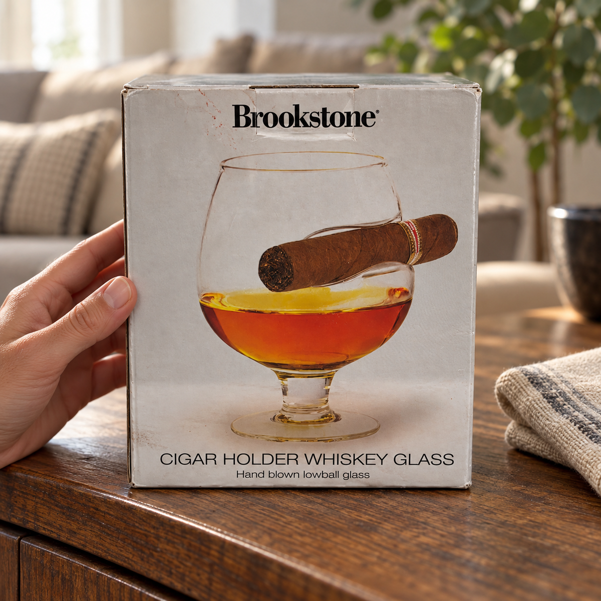AI-created lifestyle illustration of the worn Brookstone whiskey-glass retail package on a sideboard with an adult hand; setting props not included
