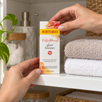 Adult hands placing Burt's Bees Truly Glowing Glow Booster carton on a cabinet shelf, AI-assisted illustration