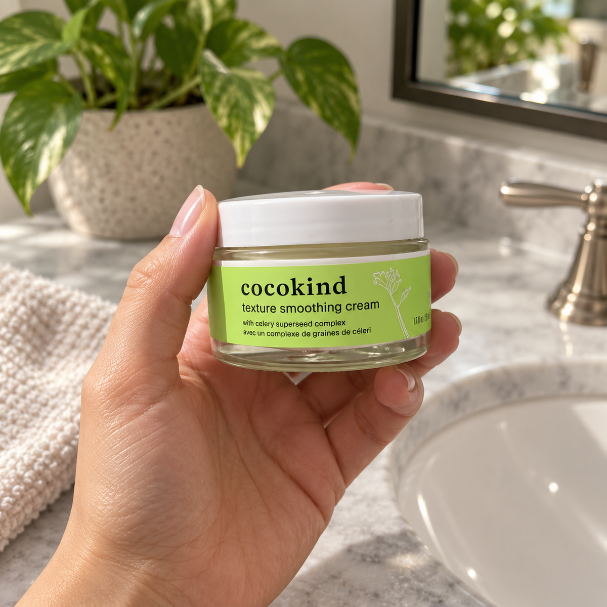 An adult holding a closed Cocokind Texture Smoothing Cream jar beside a bathroom sink