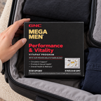 A hand arranging the closed GNC Mega Men Performance & Vitality carton beside clothing in a suitcase.