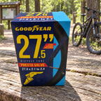 Goodyear 27.5 x 2.1–2.4 Presta valve bicycle tube package on a picnic bench at a wooded cycling trailhead; AI-staged lifestyle scene.