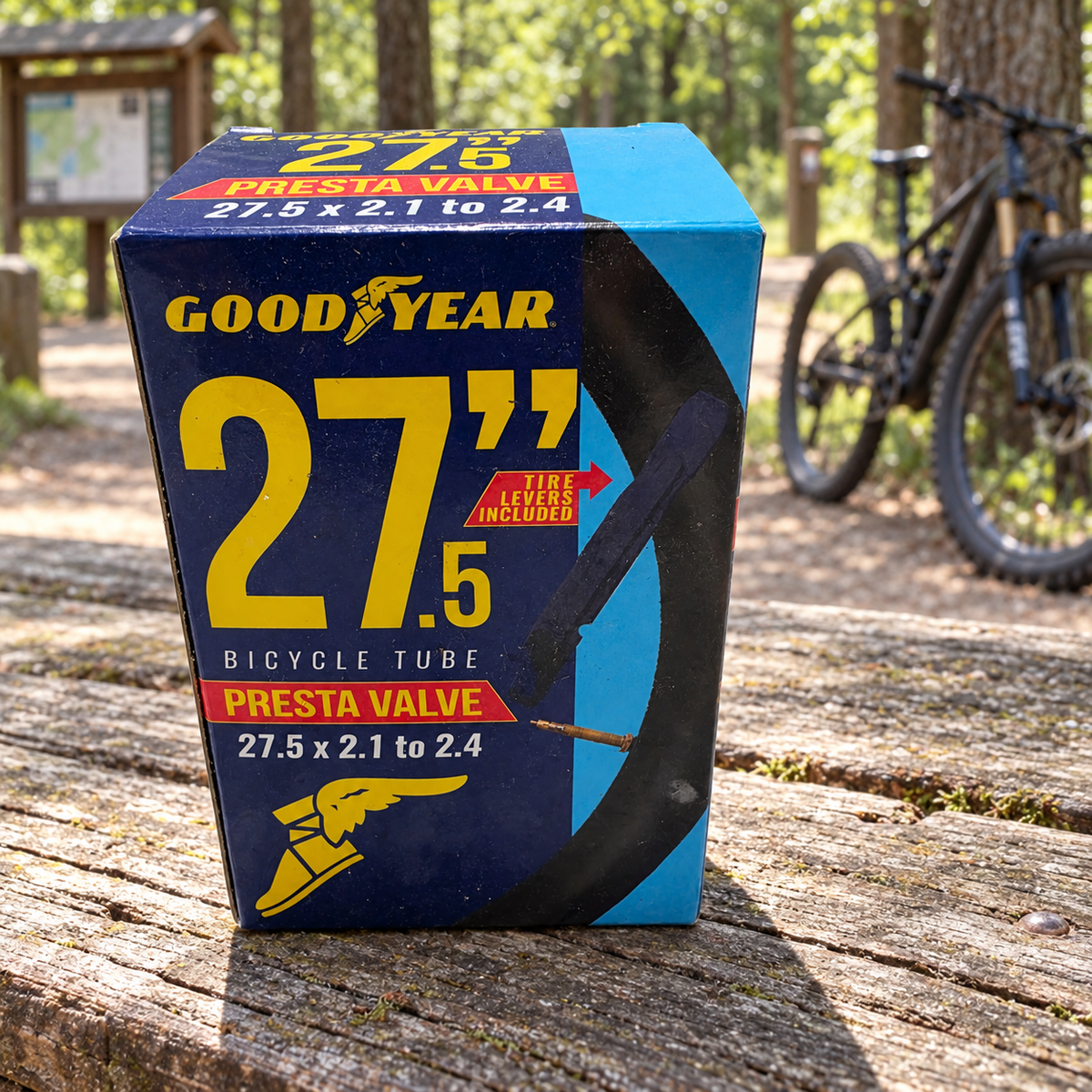 Goodyear 27.5 x 2.1–2.4 Presta valve bicycle tube package on a picnic bench at a wooded cycling trailhead; AI-staged lifestyle scene.