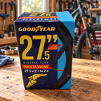 Goodyear 27.5 x 2.1–2.4 Presta valve bicycle tube retail package on a bicycle repair workbench; AI-staged lifestyle scene.
