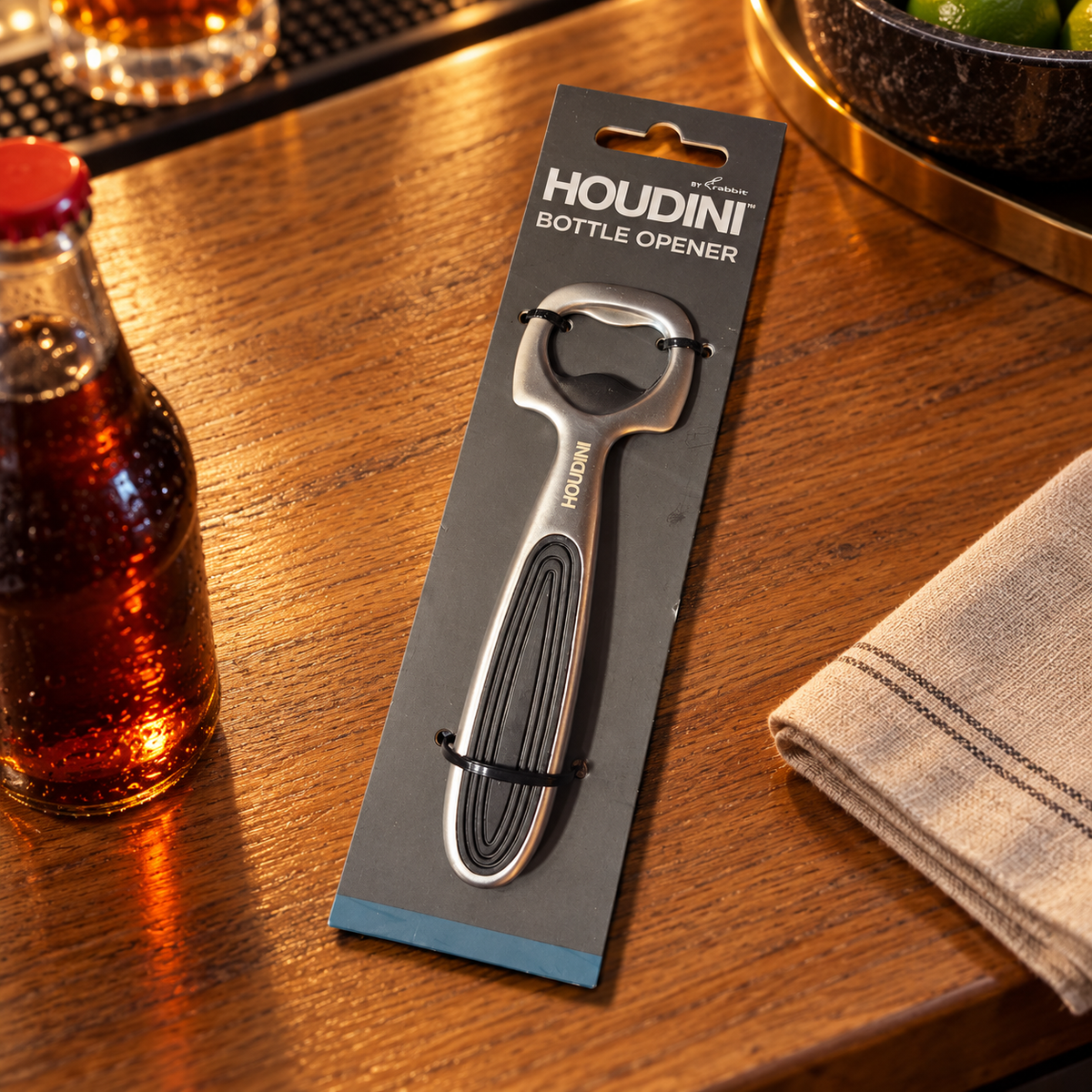 Illustrative packaged Houdini bottle opener on an oak bar counter; soda bottle and napkin not included.
