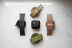 Flat lay of multiple WITHit smartwatch replacement bands in black silicone, rose gold mesh, tan perlon, and green elastic on white marble surface