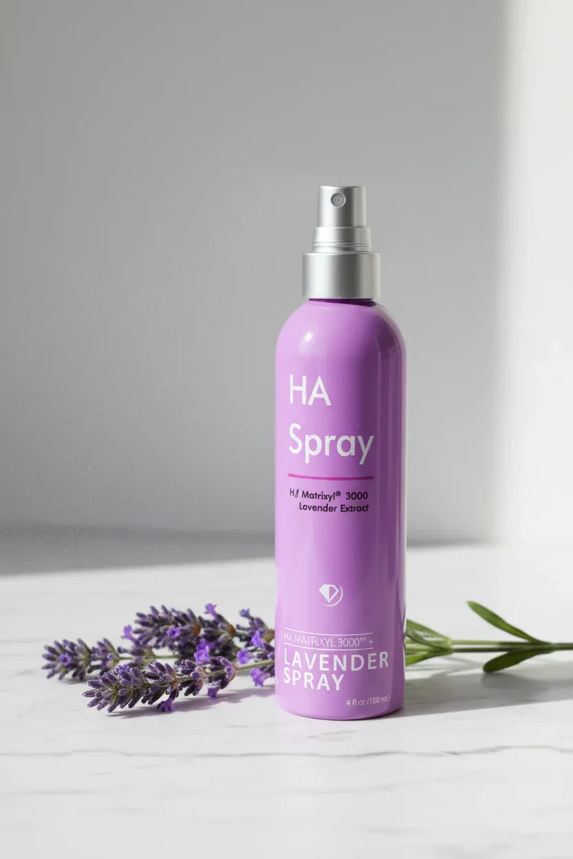 Fresh purple lavender branch next to a purple hydrating facial mist spray bottle on white marble in soft natural lighting