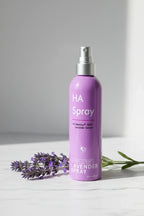 Fresh purple lavender branch next to a purple hydrating facial mist spray bottle on white marble in soft natural lighting