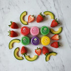 Top-down flat lay of Noro lip balm jars surrounded by fresh strawberries and avocado slices on marble