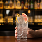 Close-up of a hand holding a tall crystal longdrink glass with citrus garnish and ice