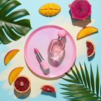 Lifestyle image — pink perfume spray bottle on bright vanity tray beside lipstick, tropical fruit slices and green leaves, colorful flatlay beauty for visual context only