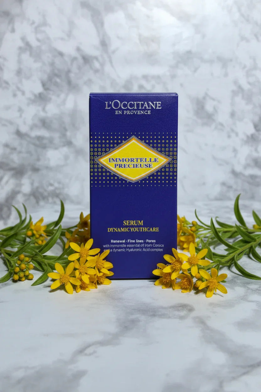 Minimalist display of botanical face serum surrounded by fresh yellow wildflowers and green leaves on white marble