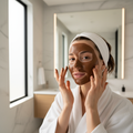 Young woman with glowing skin applying dark brown clay mud mask to her face in a bright modern bathroom