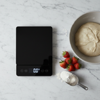 Lifestyle — sleek black glass digital kitchen scale on white marble counter beside mixing bowl, fresh strawberries, and flour scoop — clean modern baking flatlay editorial for visual context only