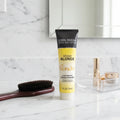 John Frieda Sheer Blonde Go Blonder Lightening Conditioner tube on white marble bathroom vanity next to wooden paddle brush and organized cosmetics tray