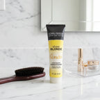 John Frieda Sheer Blonde Go Blonder Lightening Conditioner tube on white marble bathroom vanity next to wooden paddle brush and organized cosmetics tray