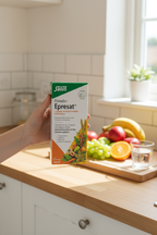 Salus Floradix Epresat Liquid Multivitamin held in hand in bright kitchen with fresh fruit in background.