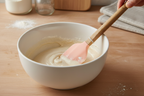 Close-up of peach silicone spatula scraping flour batter in a white ceramic mixing bowl