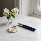 Lifestyle image — slim navy blue rollerball perfume wand on white marble vanity beside white jasmine and pearl bracelet, elegant editorial for visual context only
