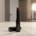 Mary Kay Lip Suede Lipstick in Mulberry Muse — black gloss tube with bullet extended showing rich berry-purple shade — standing upright on a white marble vanity with soft warm lighting and a blurred mirror in the background