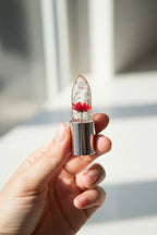 In-hand close-up of Firstfly transparent lipstick revealing gold foil and embedded dried red flower