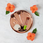 Mary Kay Dual Lip Glaze Warm Coral clear squeeze tube with green cap on a rose-gold tray with makeup brushes, a small mirror, and tropical orange flowers on white marble — feminine beauty flat-lay