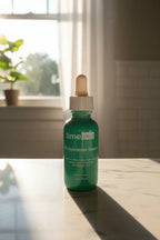 Small dark green glass skincare dropper bottle catching warm morning sunlight through a bright bathroom window on white marble