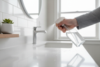 Hand spraying a clear cleaning bottle onto a white quartz sink edge in a bright modern bathroom