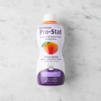 Overhead flat lay of Nutricia Pro-Stat liquid protein medical food bottle on white marble surface, bright even studio lighting