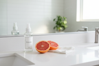 Minimalist white bathroom countertop with cleaning spray bottle, fresh-cut grapefruit halves, and folded white cloth