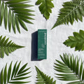 Overhead flat lay of Cocokind Chlorophyll Discoloration Serum dark green box on white linen with fresh green botanical leaves
