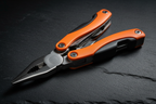Macro close-up of Adventure Is Out There 11-in-1 orange multi-tool with pliers extended on dark slate surface