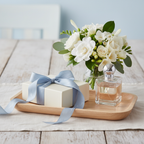 Lifestyle image — ivory gift box with blue ribbon beside clear perfume bottle and white flowers on wood vanity tray, gifting editorial for visual context only