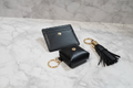Collection XIIX 3-piece black faux leather SLG set flat lay on marble — slim card holder snapped headphone case and gold-tone tassel keychain gift set