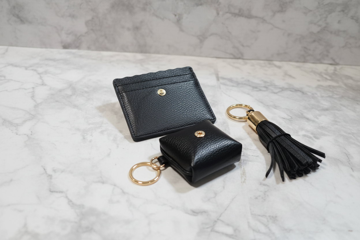 Collection XIIX 3-piece black faux leather SLG set flat lay on marble — slim card holder snapped headphone case and gold-tone tassel keychain gift set