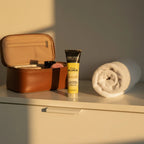 John Frieda Sheer Blonde Go Blonder Lightening Conditioner tube on minimalist vanity dresser next to open premium leather travel case and rolled white hand towel in golden hour sunlight