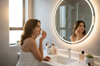 Woman applying liquid foundation at vanity mirror with glowing luminous skin in soft natural light